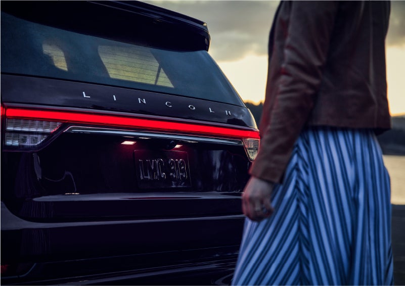 A person is shown near the rear of a 2023 Lincoln Aviator® SUV as the Lincoln Embrace illuminates the rear lights | Rydell Lincoln in Independence IA
