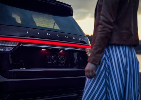 A person is shown near the rear of a 2024 Lincoln Aviator® SUV as the Lincoln Embrace illuminates the rear lights | Rydell Lincoln in Independence IA