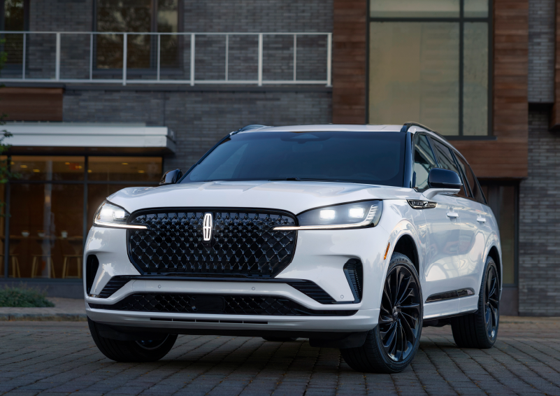 A white 2025 Lincoln Aviator® SUV is shown parked outside a modern building with gray brick and wood accents. | Rydell Lincoln in Independence IA