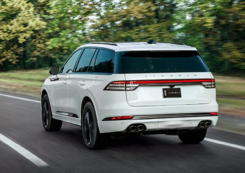 A white 2026 Lincoln Aviator® SUV is being driven away down a road lined with colorful trees. | Rydell Lincoln in Independence IA
